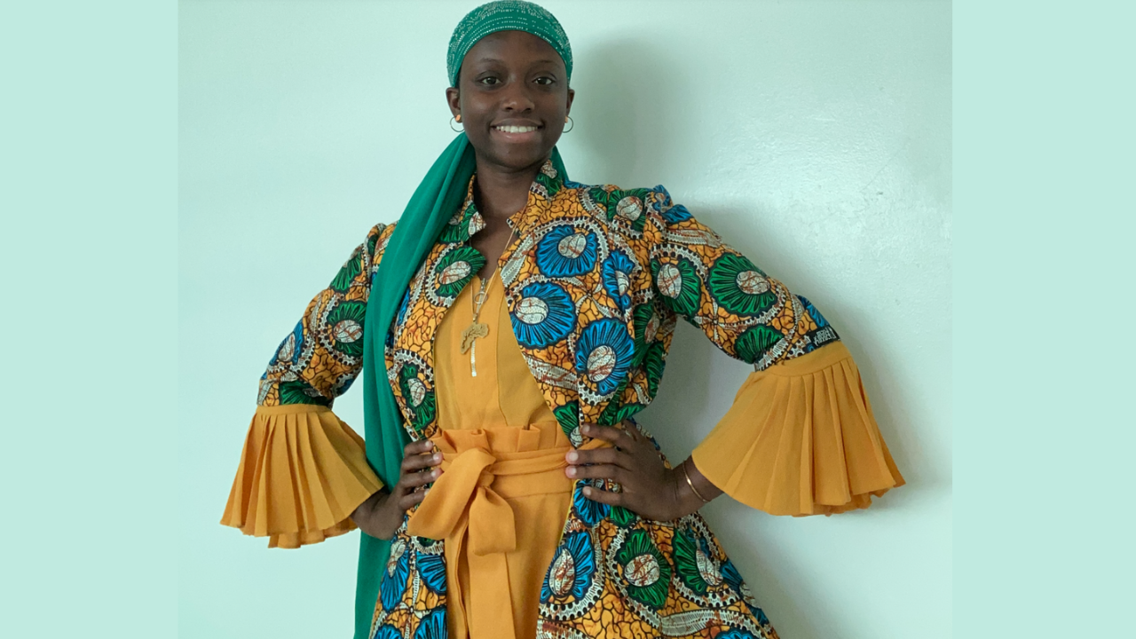 portrait of Alimah Kasumu wearing green and yellow dress standing in front of mint green wall